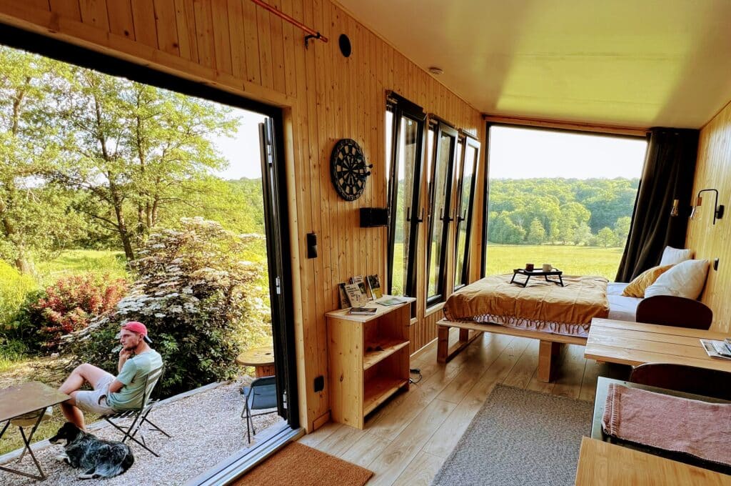 Vue de l'intérieur de la tiny house la grange avec une homme caressant un chien sur la terrasse donnant sur la nture ensoleillé. De grande ouverture permette de voir la nature totale